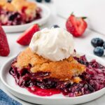 berry cobbler on a white plate with vanilla ice cream on top. berries and a baking dish in the background.