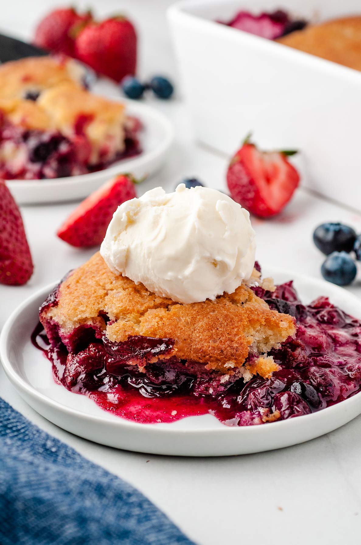 berry cobbler on a white plate with vanilla ice cream on top. berries and a baking dish in the background.