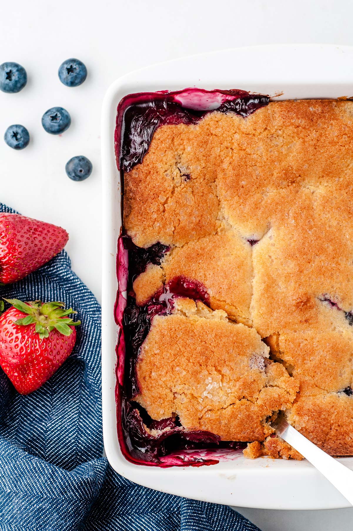 half of a white baking dish with a baked berry cobbler. blueberries, strawberries, and a blue towel on the side. 