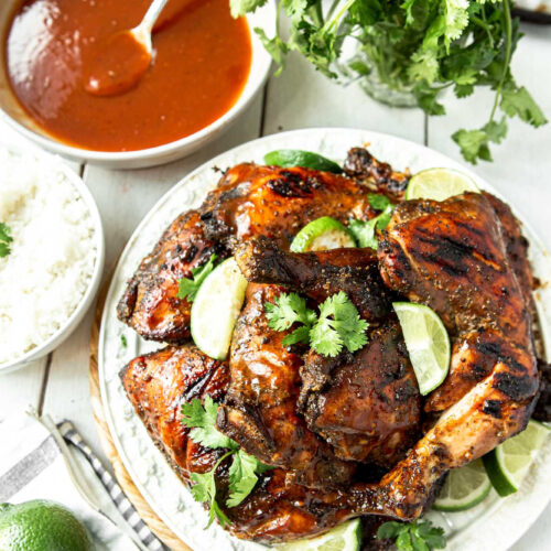 Jamaican jerk chicken on a white plate with cilantro and lime wedges. A bowl of red sauce with a spoon and more cilantro and lime on the side.