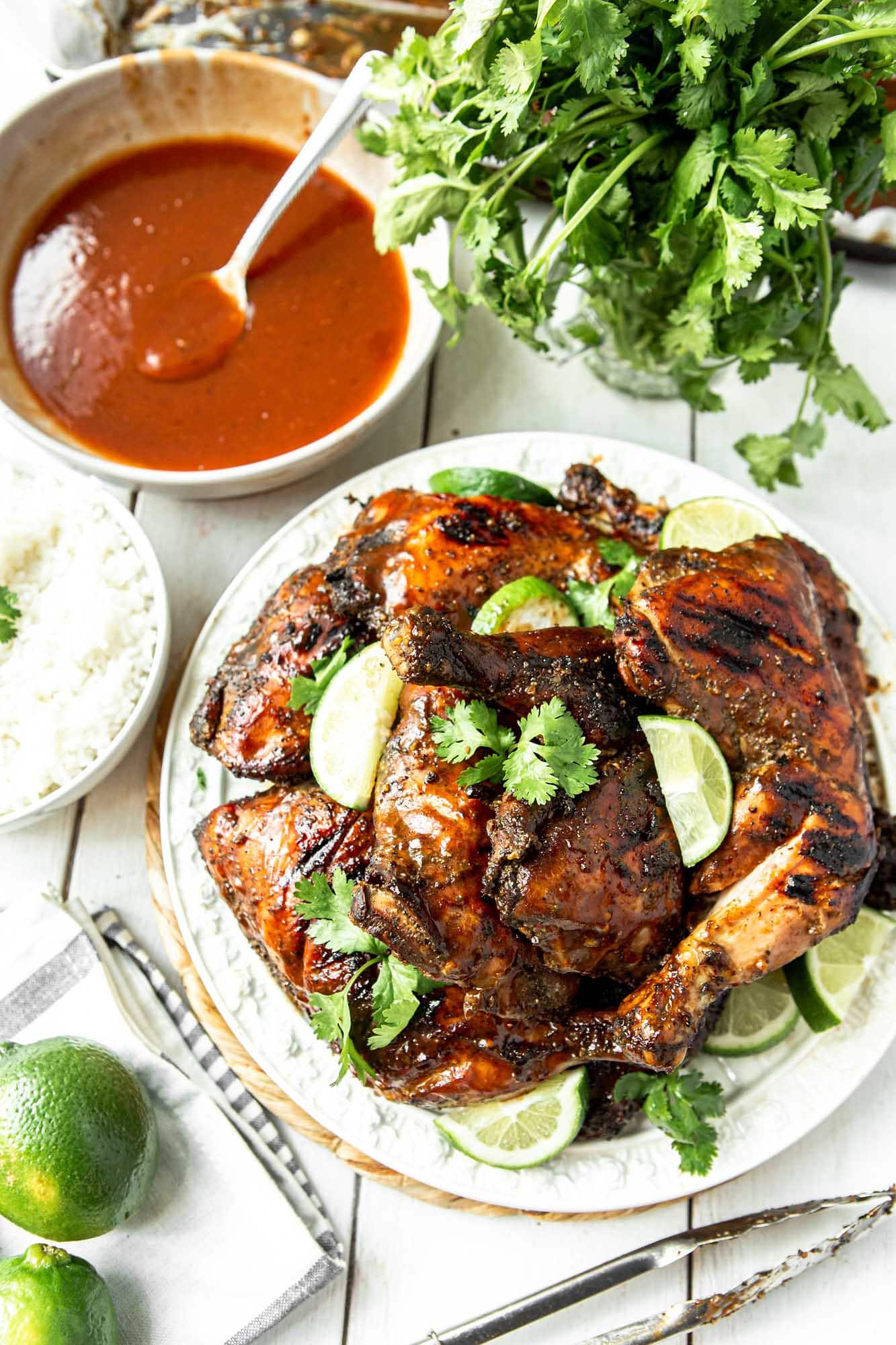 Jamaican jerk chicken on a white plate with cilantro and lime wedges. A bowl of red sauce with a spoon and more cilantro and lime on the side. 