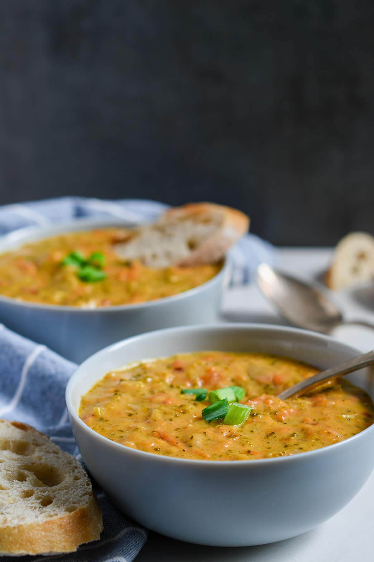 two bowls filled with broccoli and cheese soup with scallions on top, bread on the side, a blue and white towel, and a dark backdrop. 