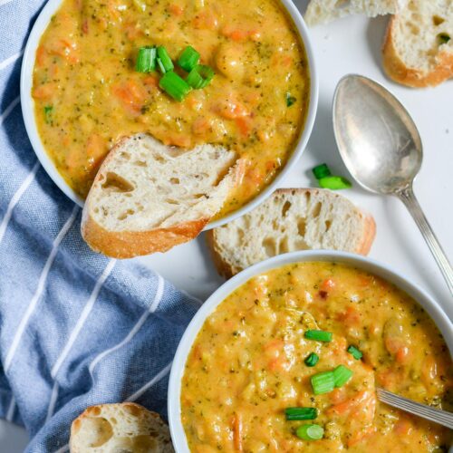 two white bowls with broccoli cheddar soup, slices of bread inside one bowl and on the side. A spoon on the side and a blue and white towel.