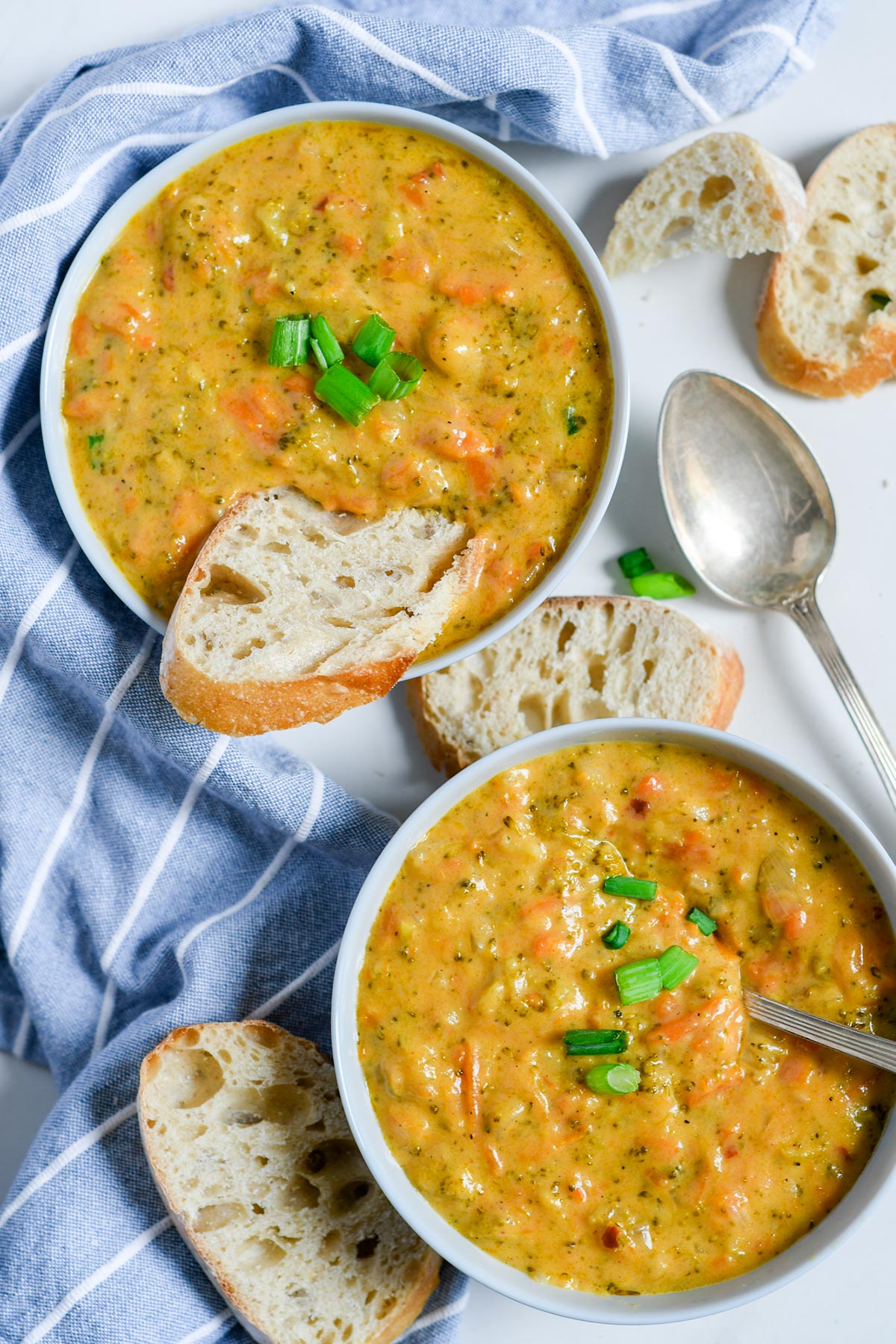 two white bowls with broccoli cheddar soup, slices of bread inside one bowl and on the side. A spoon on the side and a blue and white towel. 