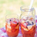 two glasses filled with sangria and fruit on top of a red and white table cloth and a pitcher with sangria in the background.