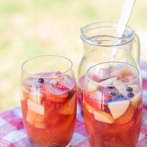 two glasses filled with sangria and fruit on top of a red and white table cloth and a pitcher with sangria in the background.