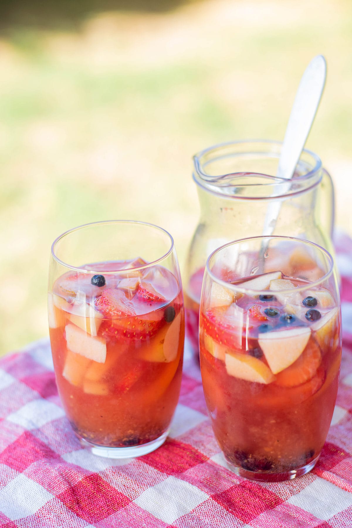 two glasses filled with sangria and fruit on top of a red and white table cloth and a pitcher with sangria in the background.