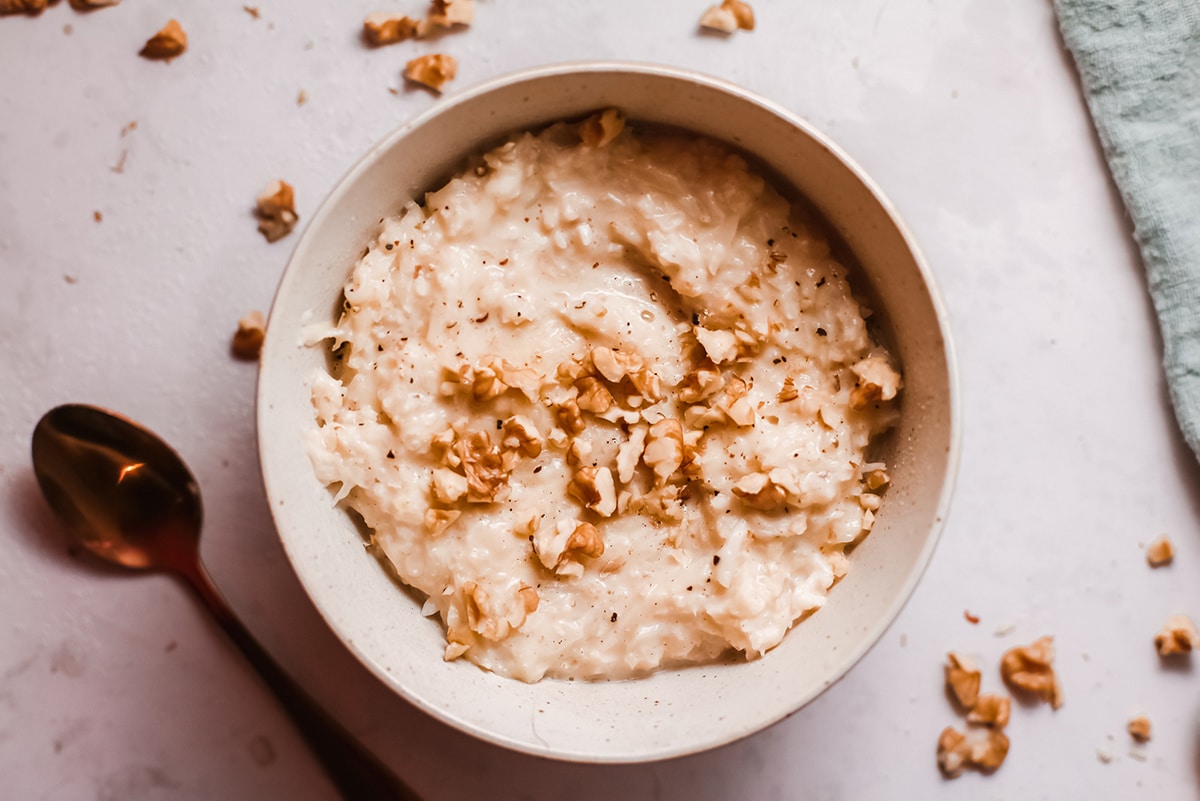 a horizontal photo of risotto in a bowl with nuts on top and on the side. A spoon on the side. 