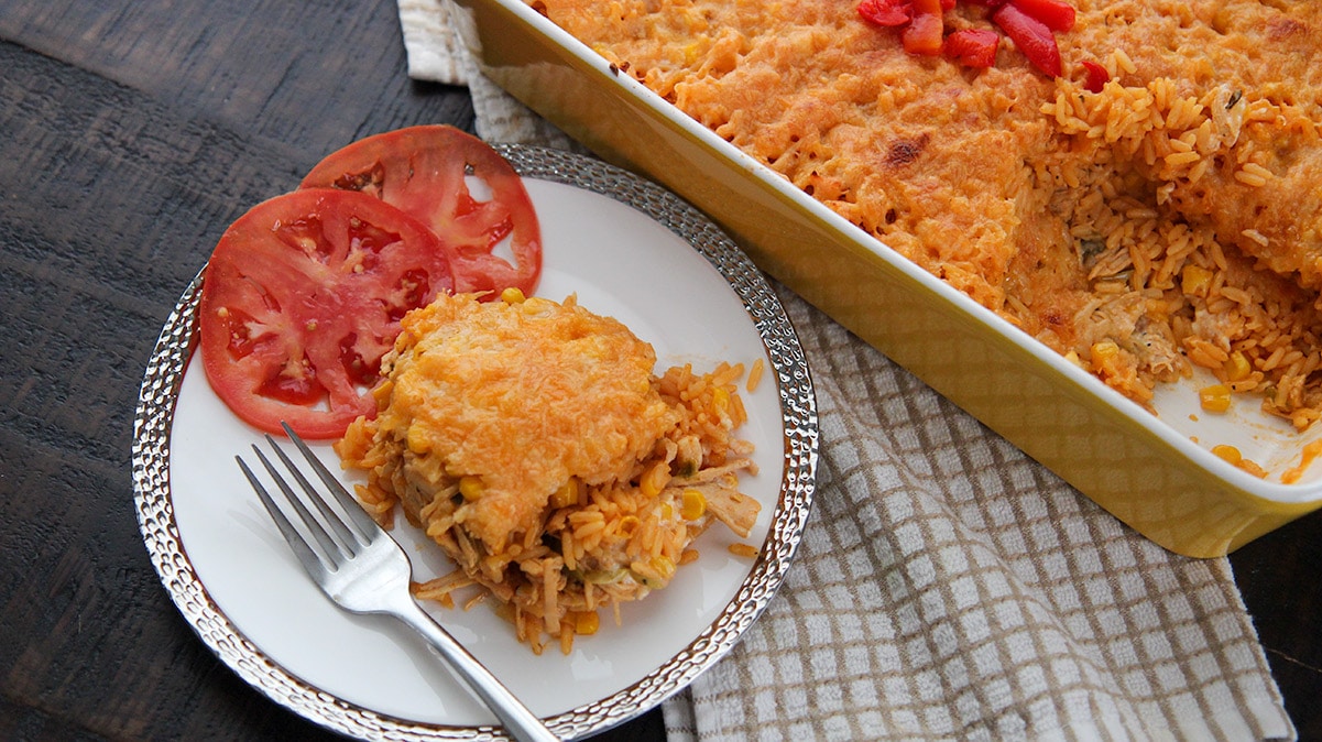 a serving of yellow rice, shredded chicken, cheese, and two tomato slices on a white plate with a fork. A yellow casserole dish in the background. 