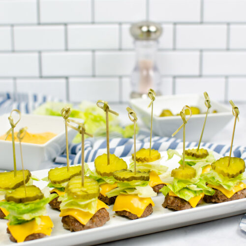 ten big mac bites with lettuce, cheese, and pickles on top of a white plate with sauce, pickles, and pepper in the background.