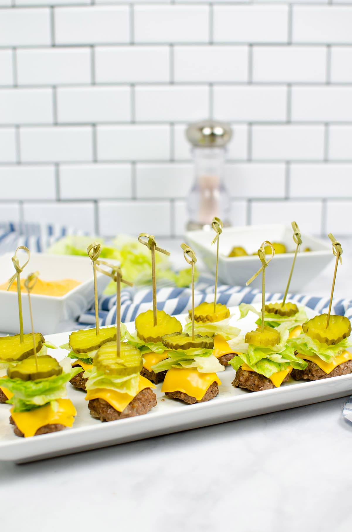 ten big mac bites with lettuce, cheese, and pickles on top of a white plate with sauce, pickles, and pepper in the background.