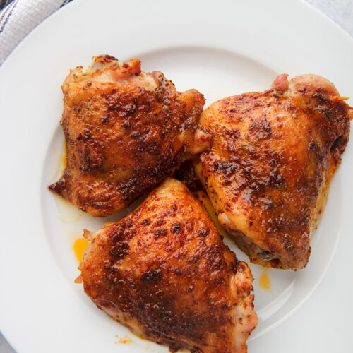 three baked chicken thighs on a white plate with a gray and white towel on the side.