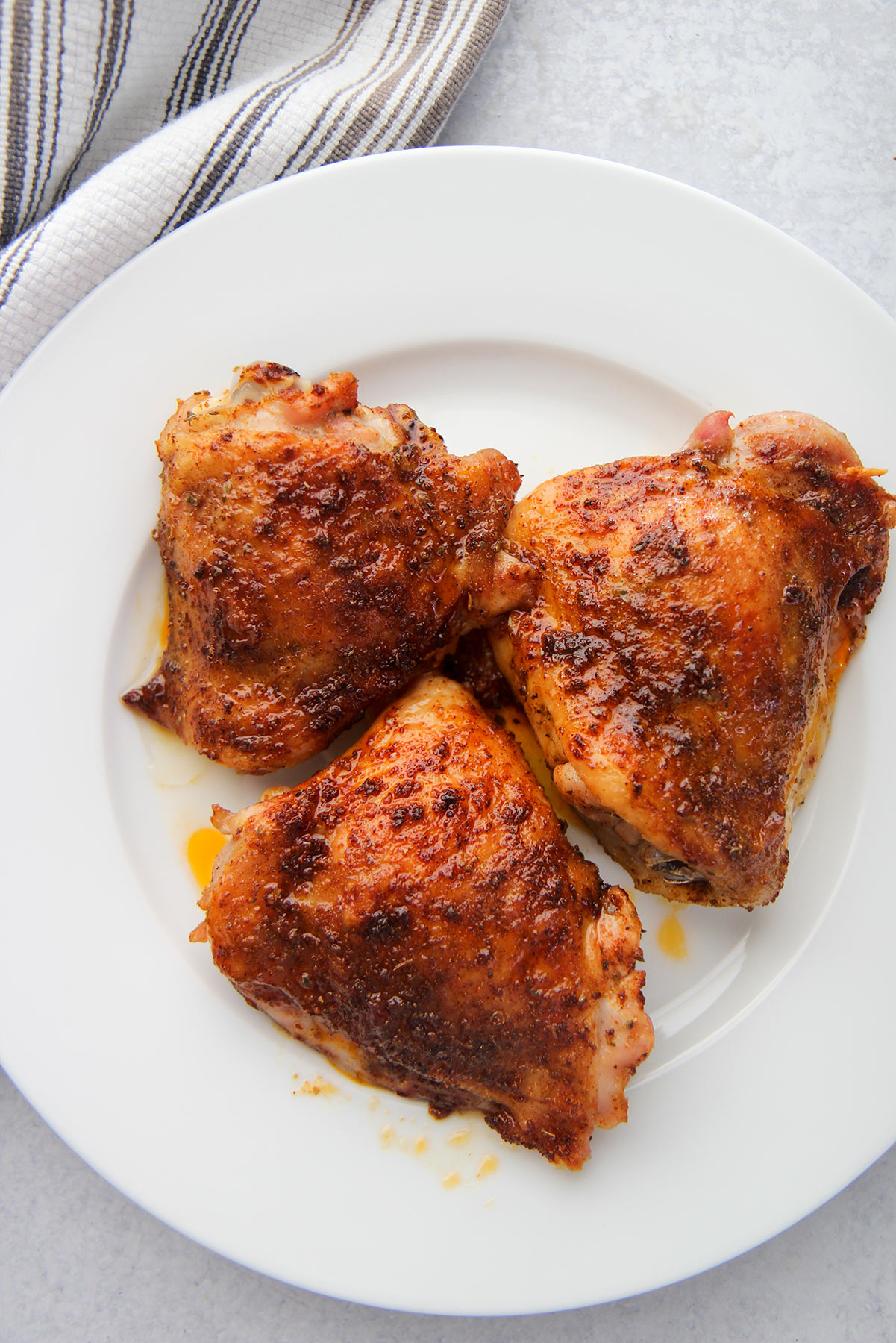 three baked chicken thighs on a white plate with a gray and white towel on the side.