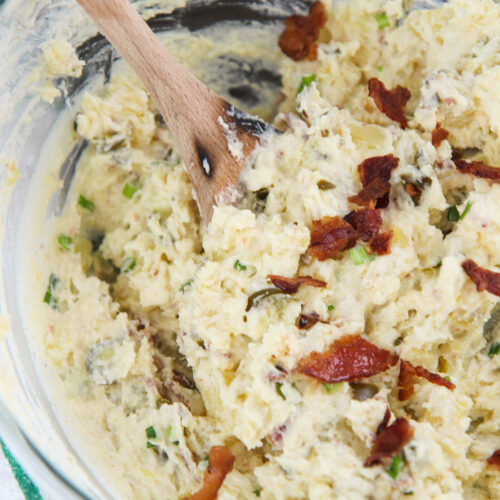 spicy potato salad in a glass bowl with bacon on top, a wooden spoon, and a green and white towel on the side.
