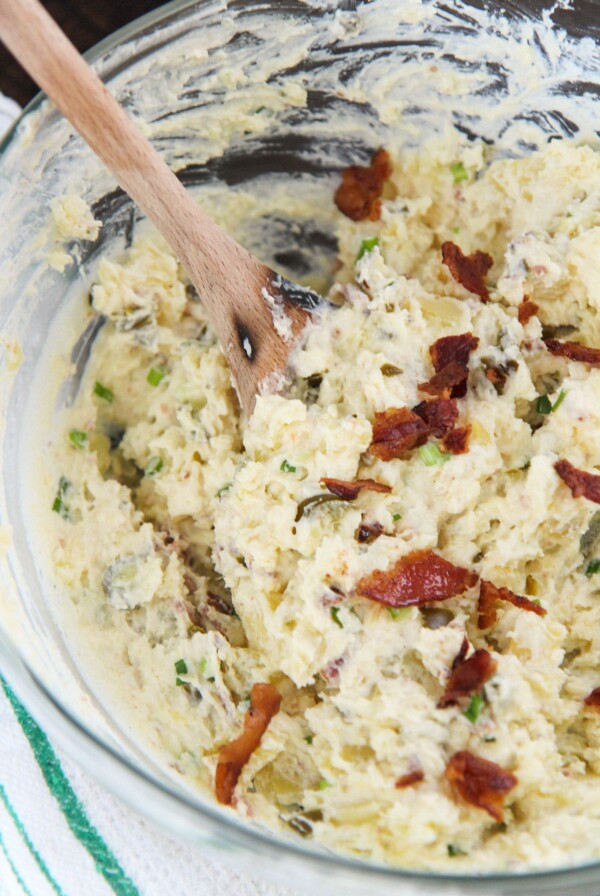 spicy potato salad in a glass bowl with bacon on top, a wooden spoon, and a green and white towel on the side.