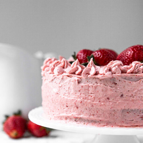 two layer fresh strawberry cake on a white cake stand with a white pitcher in the background and strawberries on the side.