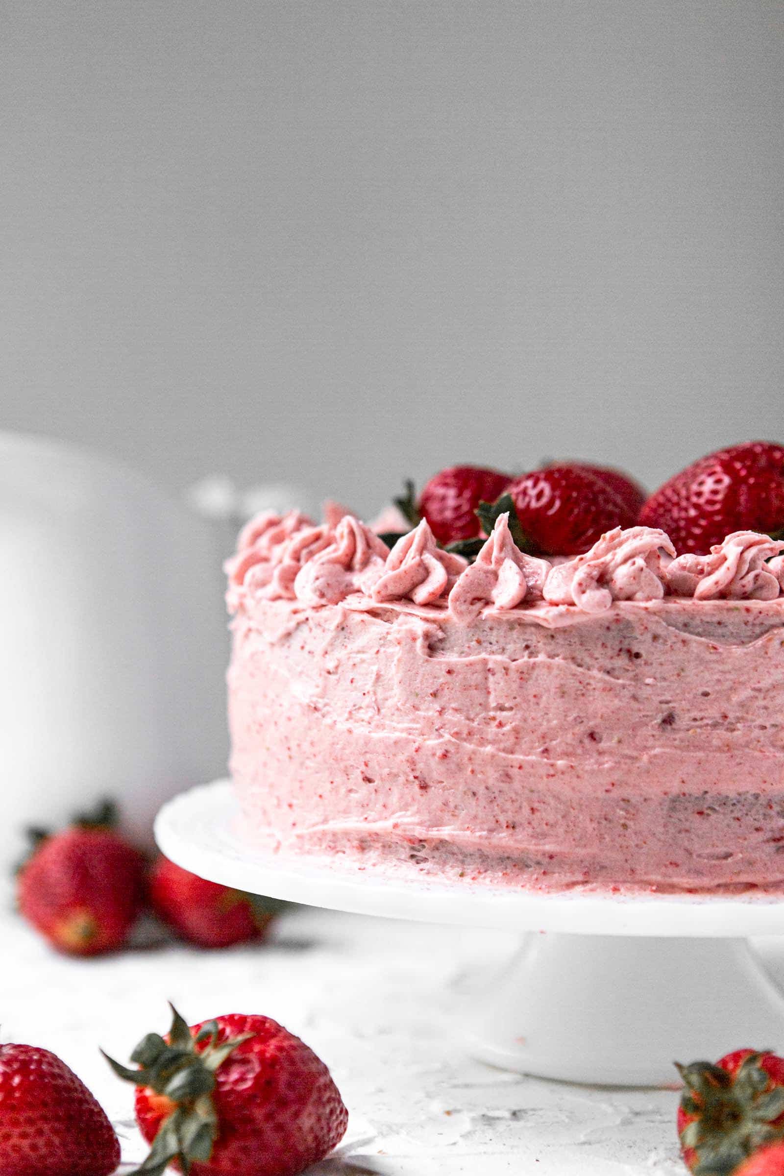 two layer fresh strawberry cake on a white cake stand with a white pitcher in the background and strawberries on the side.