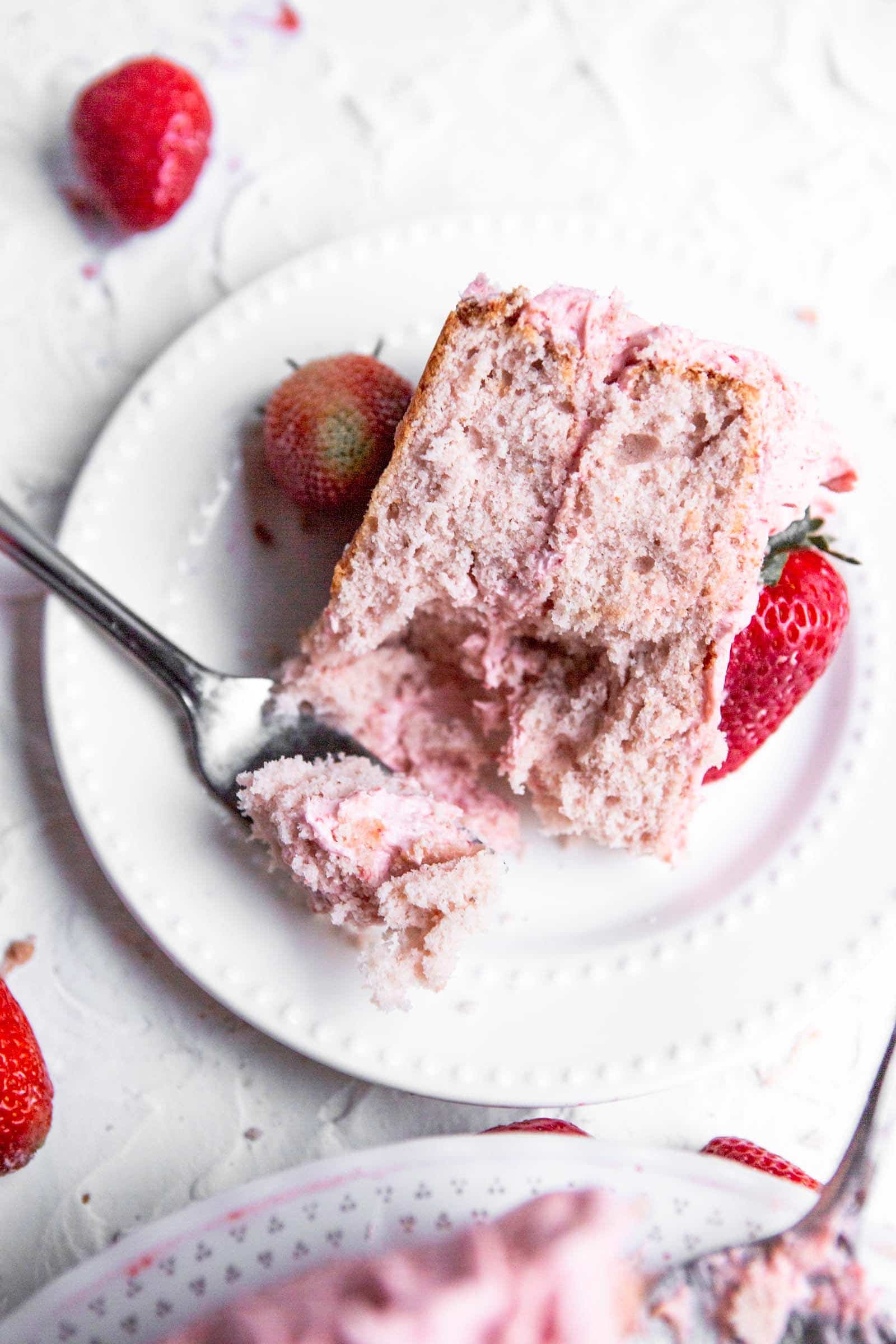 a slice of a two layer pink cake on a white plate with a fork and strawberries on the side.