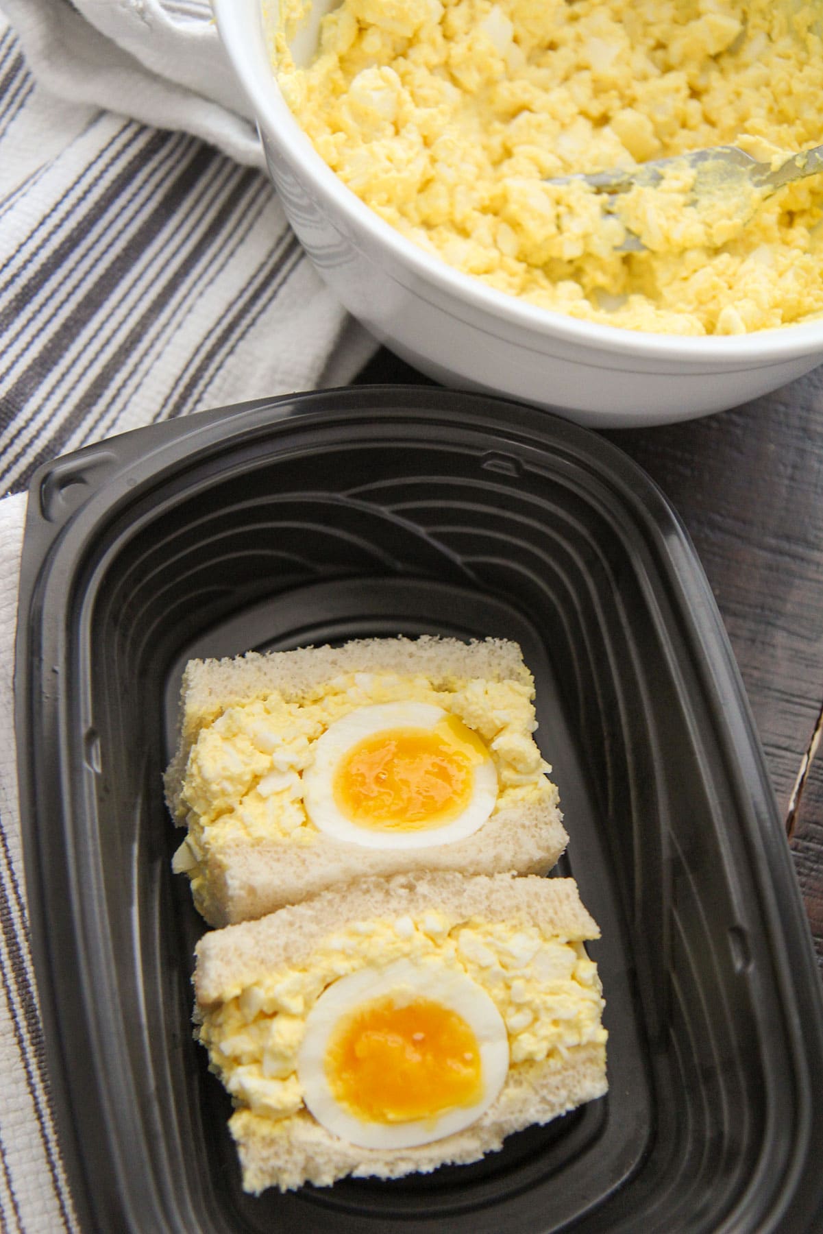 Japanese egg salad sandwiches in a black container with a bowl of egg salad in the background.