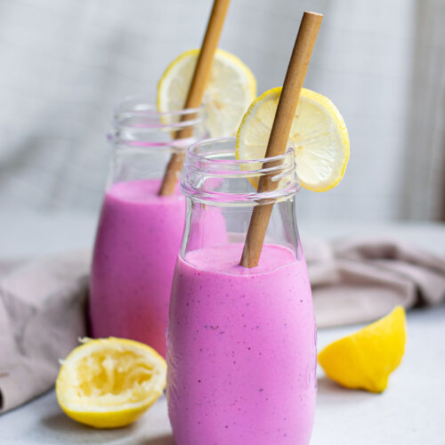 two glass jars filled 3/4s of the way with pink dragon fruit smoothie with lemons and brown straws.