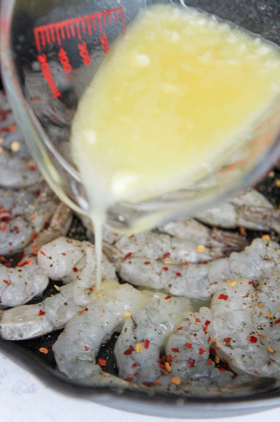 a measuring cup pouring melted butter over raw shrimp. 