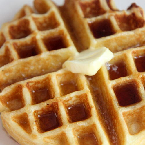 buttermilk waffle with syrup and butter on a white plate up close.