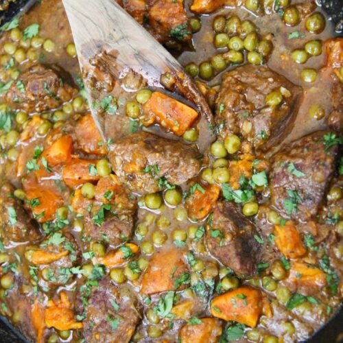 sweet potatoes, green peas, beef cubes, and a wooden spoon in a cast iron skillet.