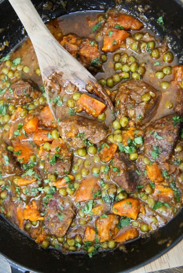 sweet potatoes, green peas, beef cubes, and a wooden spoon in a cast iron skillet.