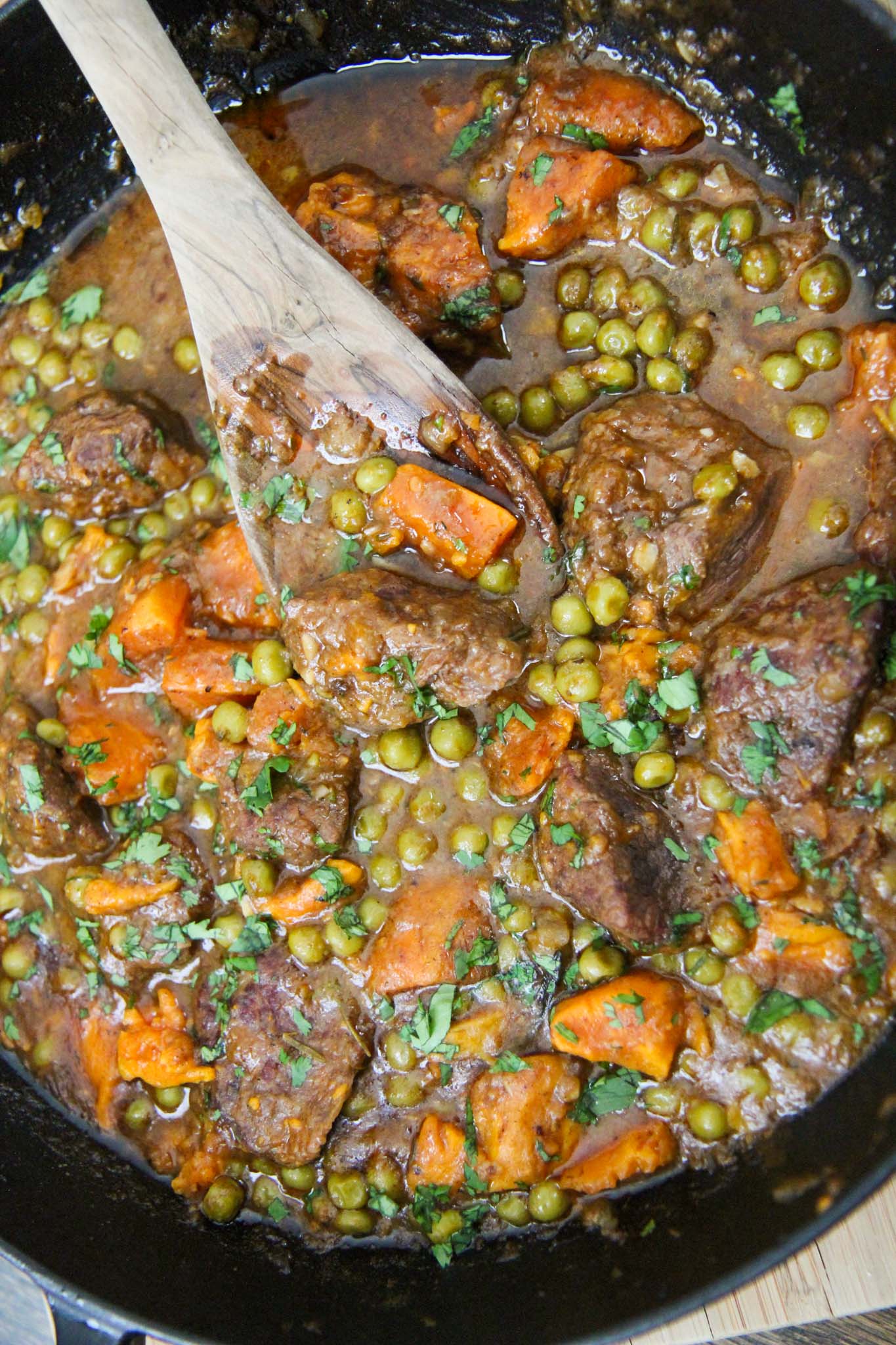 sweet potatoes, green peas, beef cubes, and a wooden spoon in a cast iron skillet. 