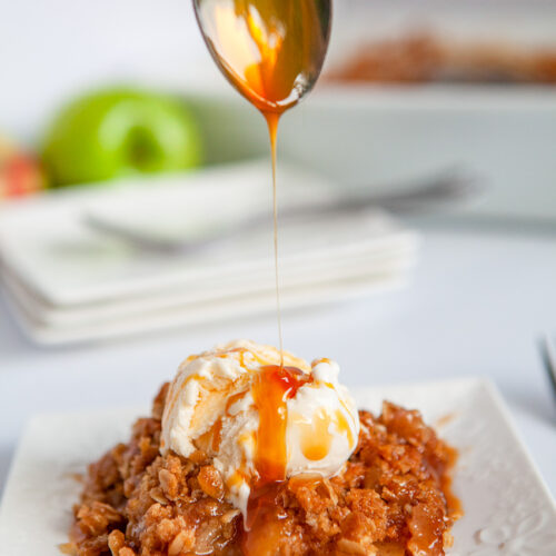 a spoon drizzling caramel sauce over apple crisp with vanilla ice cream on a white plate.