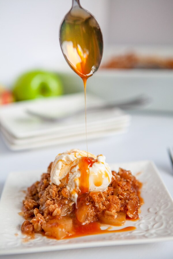 a spoon drizzling caramel sauce over apple crisp with vanilla ice cream on a white plate.