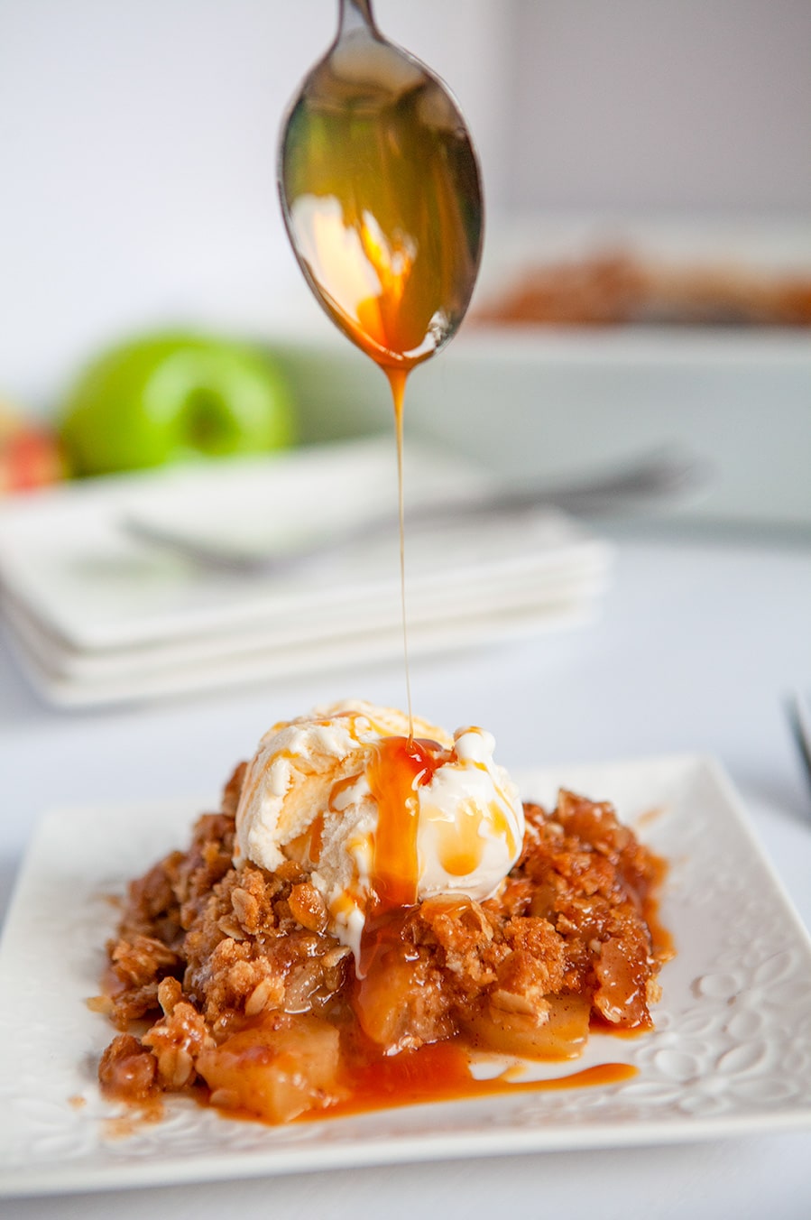 a spoon drizzling caramel sauce over apple crisp with vanilla ice cream on a white plate. 
