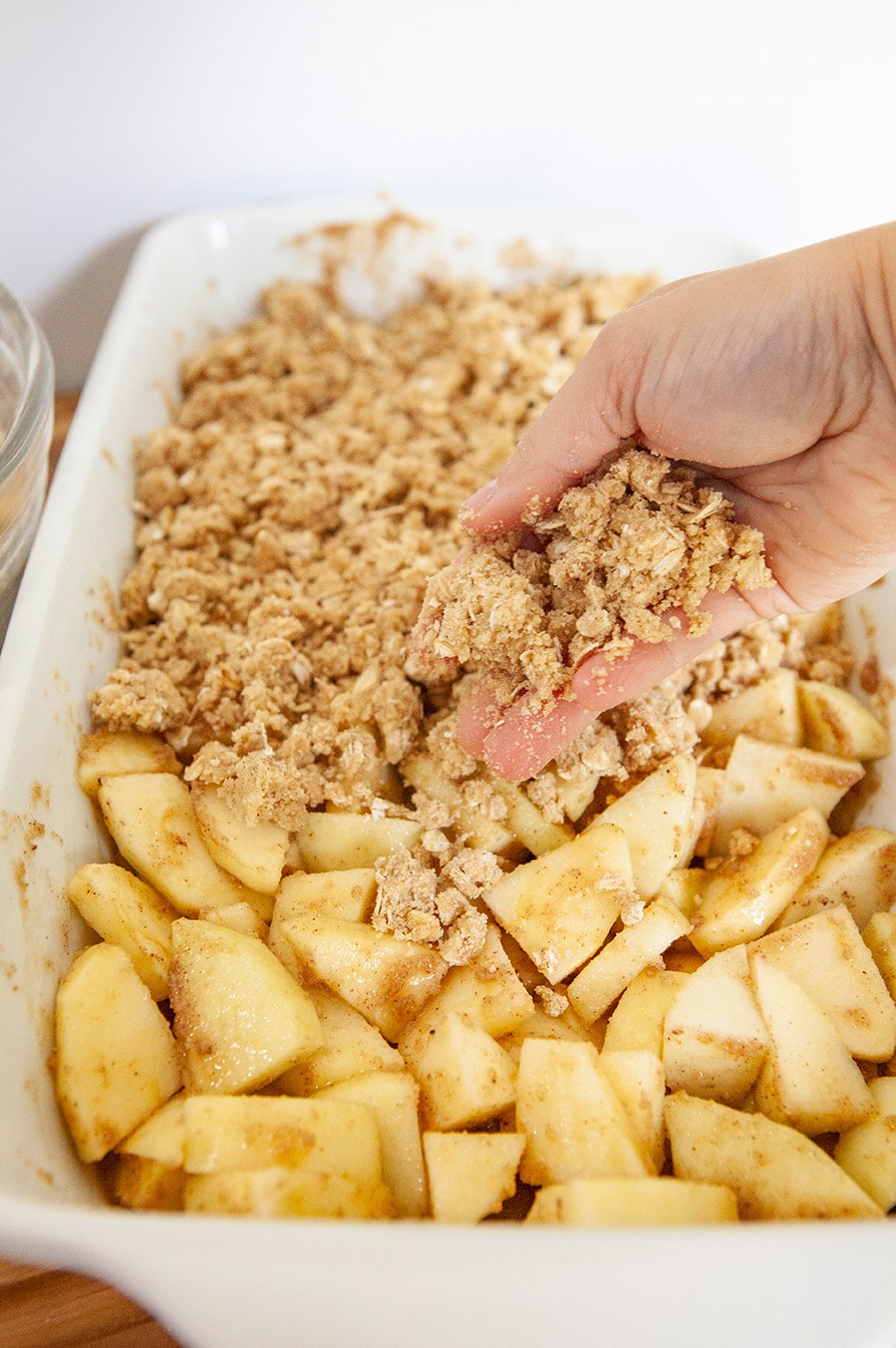a hand pouring the oat topping over apples in a white casserole dish. 
