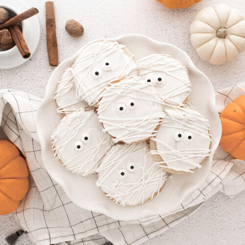 mummy cookies on a white plate with pumpkins on the side.