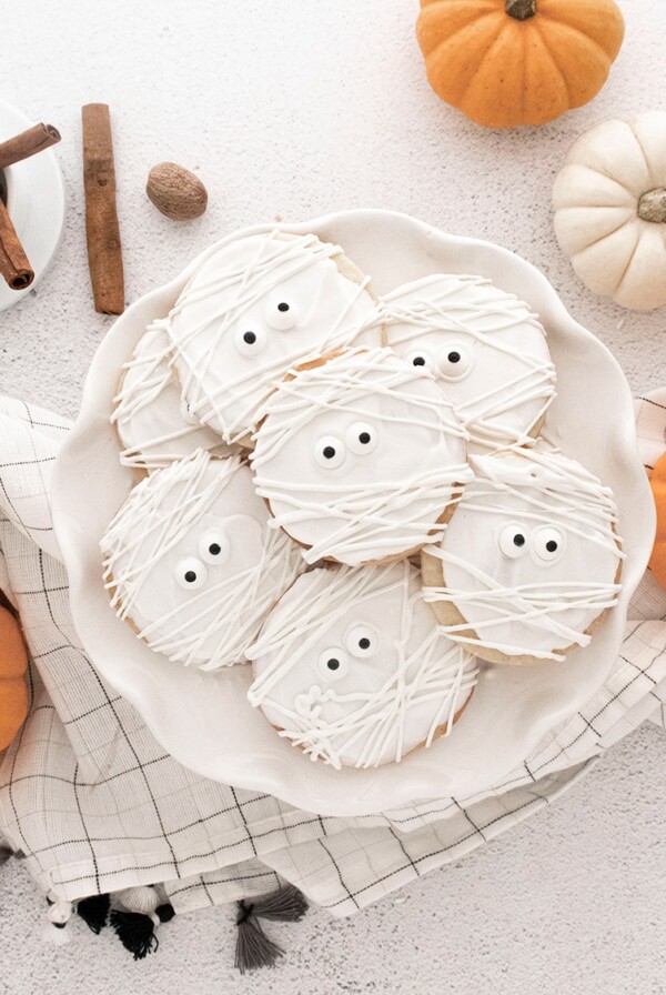 mummy cookies on a white plate with pumpkins on the side.