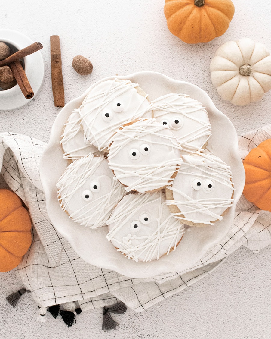 mummy cookies on a white plate with pumpkins on the side.