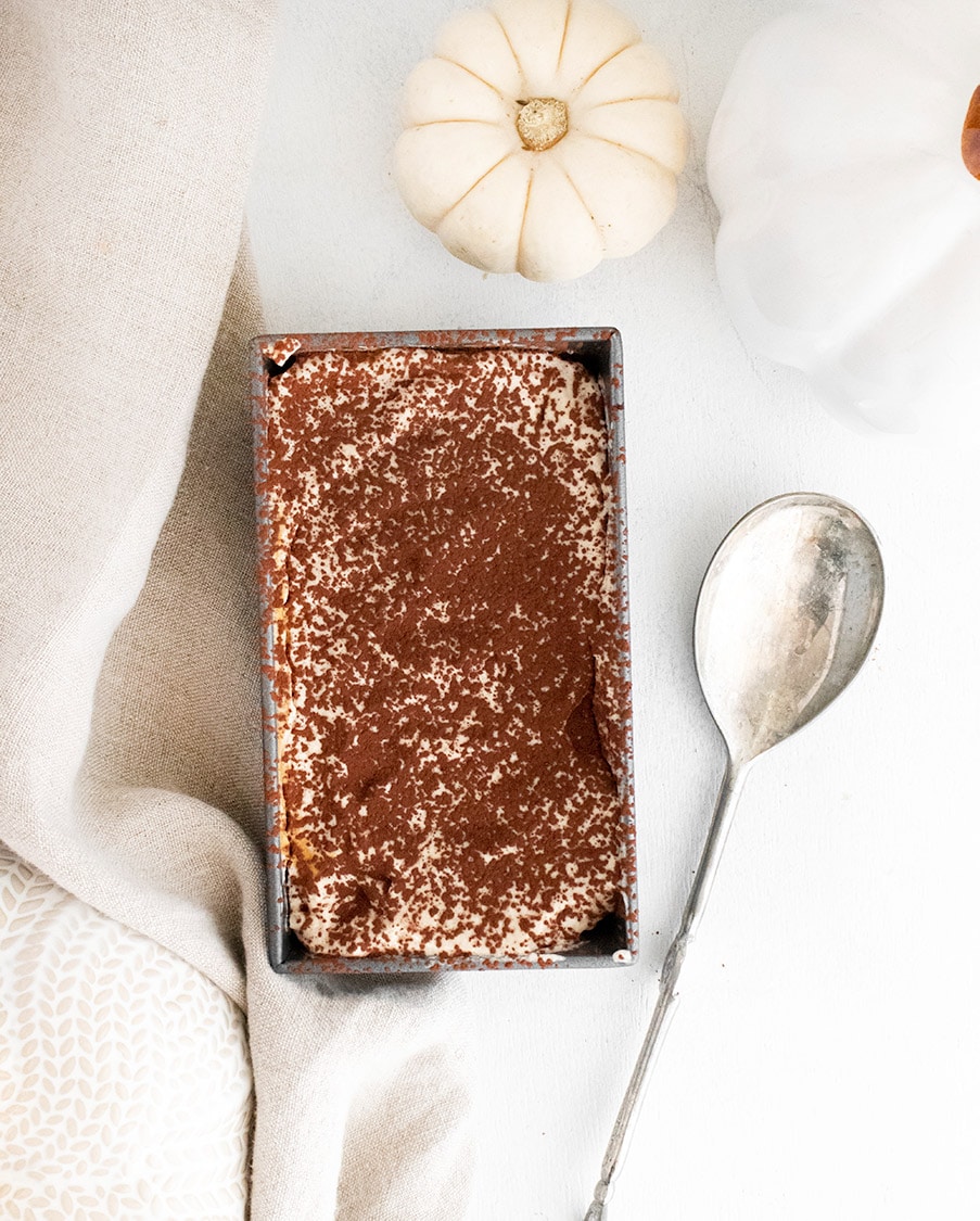 tiramisu in a loaf pan with a dark topping, a silver spoon, a light towel, and a white pumpkin on the side. 