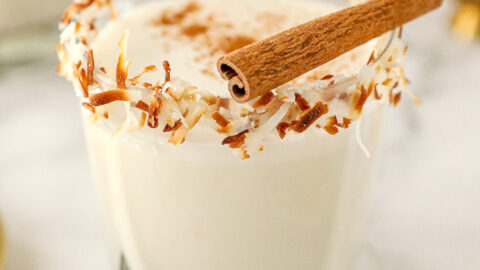 coquito in a short glass with toasted coconut on the rim and a cinnamon stick on top.
