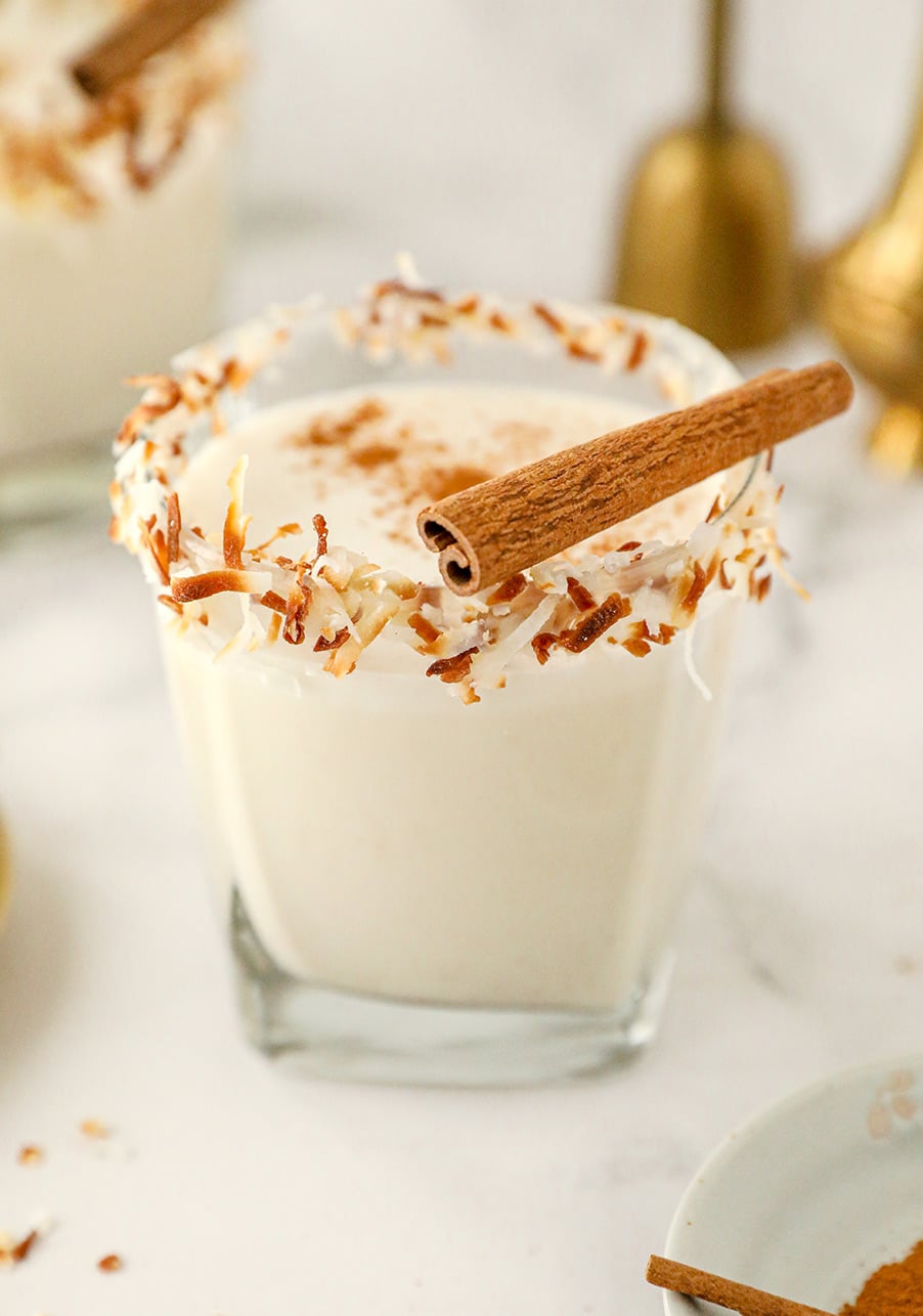 coquito in a short glass with toasted coconut on the rim and a cinnamon stick on top. 