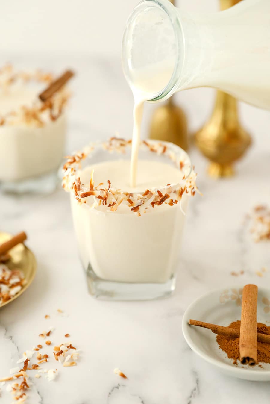 a milk jug pouring coquito into a short glass. 