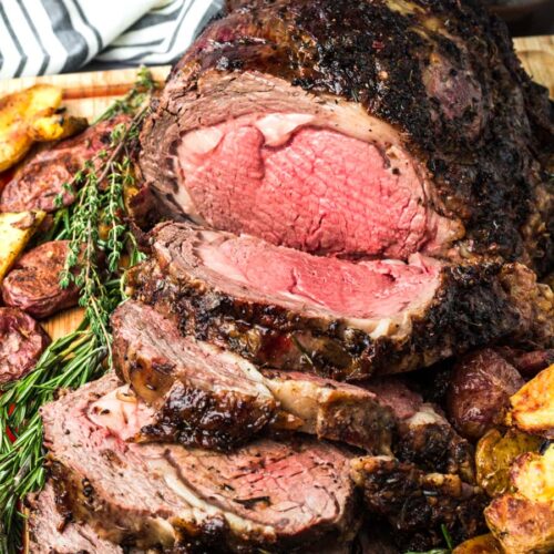 sliced prime rib medium rare on a wooden board with fresh rosemary and thyme.