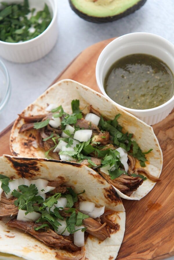 two instant pot pork carnitas tacos on a wooden board with green sauce on the side.