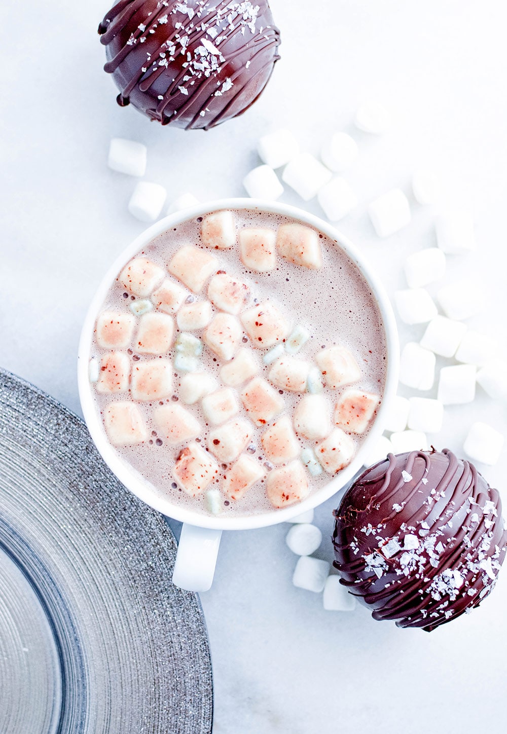 a mug filled with hot cocoa and marshmallows with two salted caramel hot cocoa bombs on the side. 