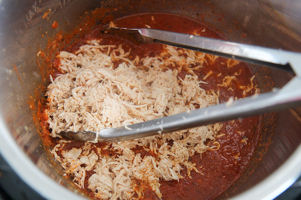 shredded white meat chicken and red sauce in the instant pot with some tongs.