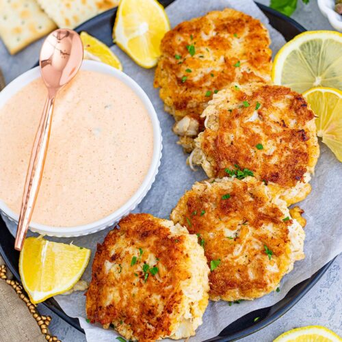 four crab cakes on a plate with a side of remoulade sauce and lemon wedges.