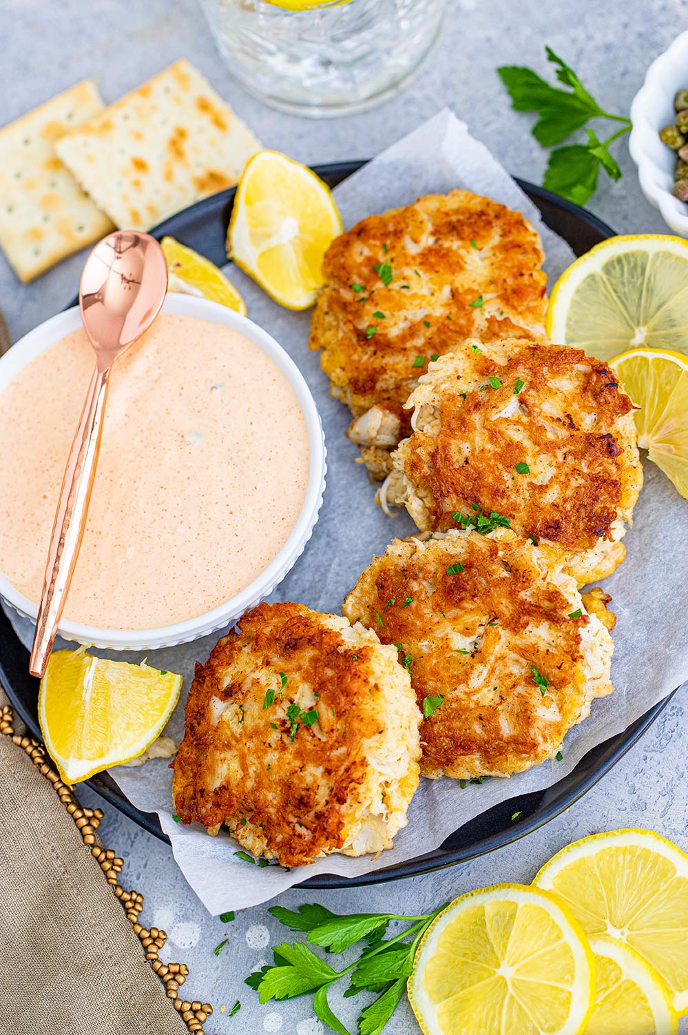 four crab cakes on a plate with a side of remoulade sauce and lemon wedges.