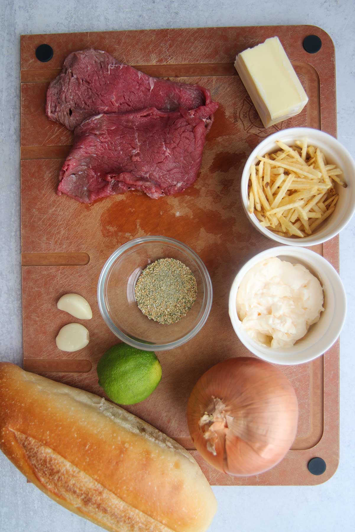 ingredients for pan con bistec. Spices in a small bowl, mayo in a bowl, steaks, lime, garlic, onion, butter, and potato straws. 