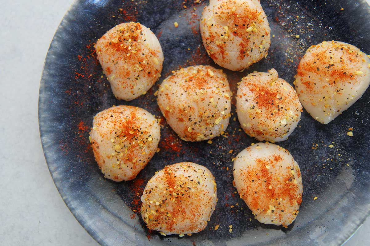 eight uncooked scallops seasoned with spices on top of a blue plate. 