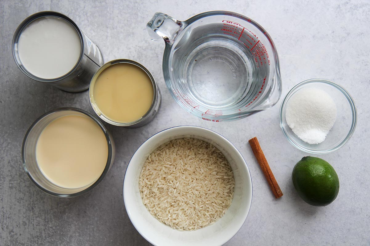 three cans of milk, water, rice in a bowl, a lime, a cinnamon stick, and sugar and salt in a small bowl. 