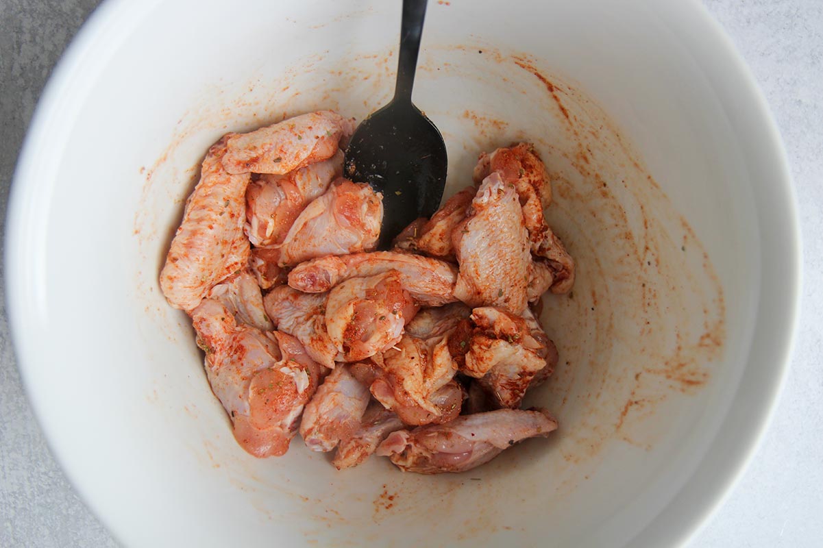 raw seasoned chicken wings in a white bowl with a black spoon.